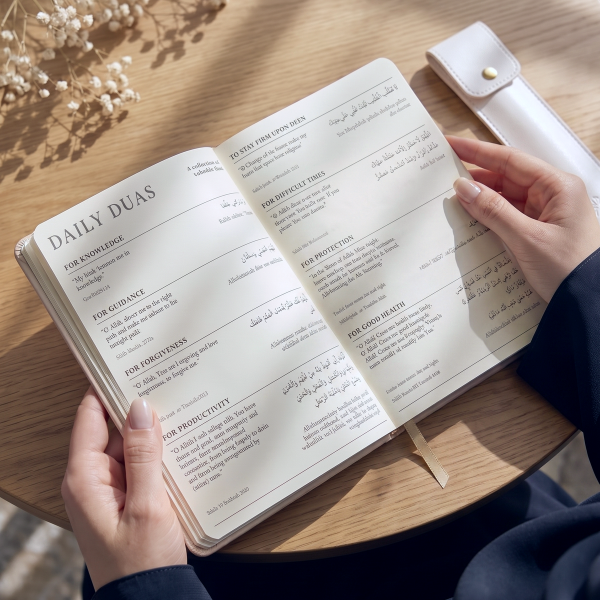 An open daily duas planner with printed prayer prompts in English and Arabic, resting on a light wood table. A person's hands are holding the book, and a white sleeve is placed beside it.