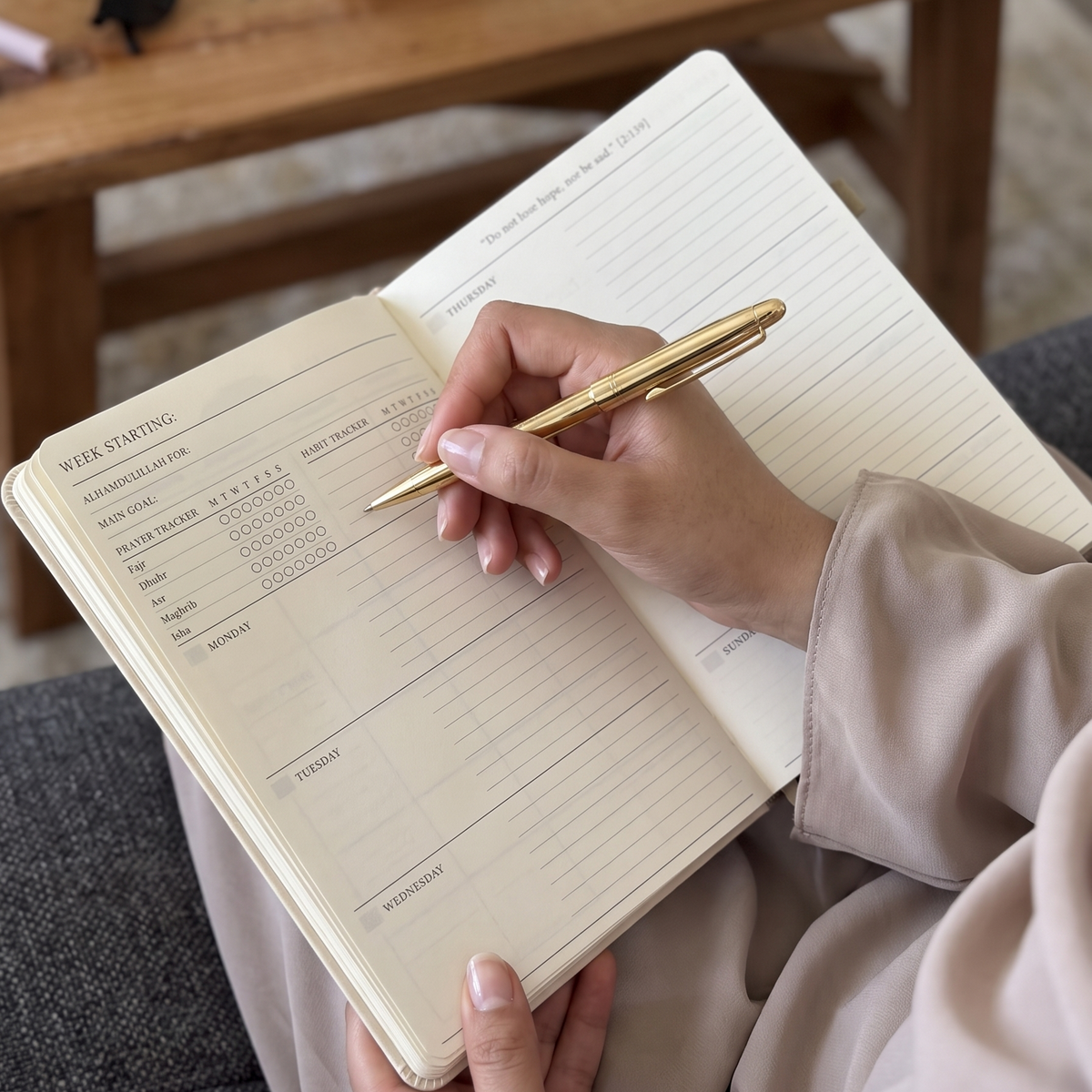 A person holding an open lined weekly planner or journal with a gold pen, seated in a cozy indoor setting with a wooden table and mug in the background.